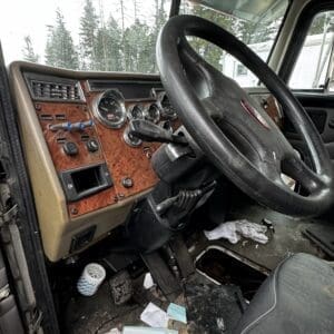Driver's-side view of a dusty, cluttered truck cabin with a large steering wheel, wood-grain dashboard, pedals, and scattered trash on the floor.