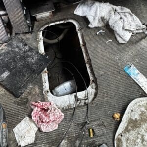 Open access panel on a dirty car floor with exposed wires and debris, including rags, a bottle, and a blue wrapper nearby (driver’s side).