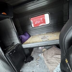 A cluttered truck sleeper cab with a wooden bed platform, purple pillow, and a small window showing a red exterior outside; scattered papers and a quilt on the floor.