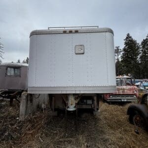 Rear view of a white box trailer raised on a hitch in a junkyard, with rusted trucks and tall trees in the background.
