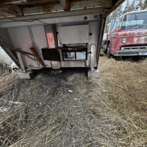 Rusty trailer underside with an open electrical panel and tangled wires; a red vintage truck is parked on dry grass nearby.