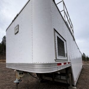 Large silver metal trailer parked on a dirt lot, with riveted panels and a side vented hatch.