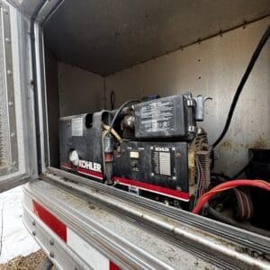 Portable generator (Kohler) mounted in a metal utility trailer, with red stripe and exposed wiring inside a weathered storage compartment.