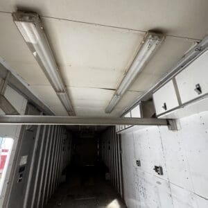 Interior of a long, empty trailer or shipping container with overhead fluorescent lights and metal walls fading to a dark rear opening.