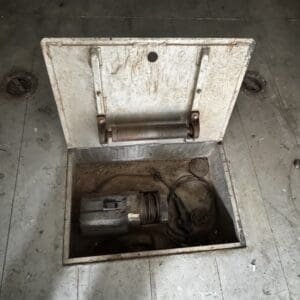 Open floor hatch reveals dusty mechanical shaft with a cylindrical motor and tangled cables inside the pit area in a dirty workshop.