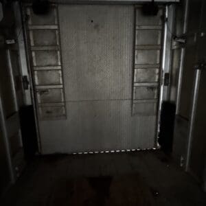 Dim, industrial metal roll-up door in a narrow corridor with ladders mounted on both sides.