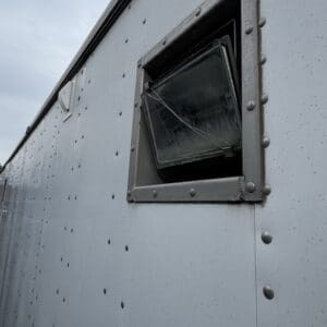 Tilted rectangular vent window on a riveted metal trailer wall, with a wire across it and cloudy sky in the background.