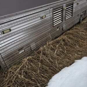 Side view of a silver aluminum trailer with vent panels, riveted siding, and small lights, next to dry grass and a patch of snow at the ground level.