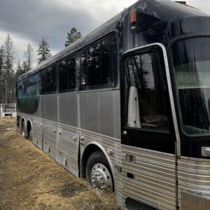 Silver and black tour bus abandoned in a dry, grassy field with flat tires and a cloudy sky overhead.