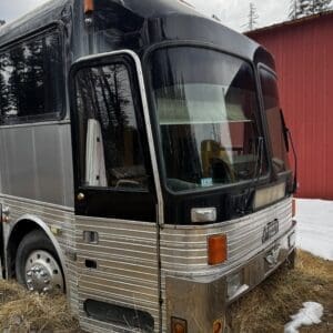 Front-three-quarters view of a silver and black tour bus parked on brown grass with patches of snow, a red barn in the background, and bare trees reflected in the windshield.