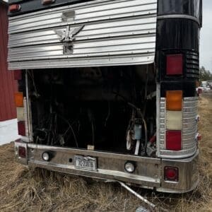 Rear of a vintage bus with its engine compartment open, exposing tangled wiring and mechanical parts, license plate TC-2387B on the bumper.