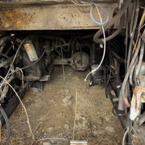 Underbelly of a dirty, rusted vehicle showing tangled hoses, exposed wiring, and a rocky dirt floor beneath.