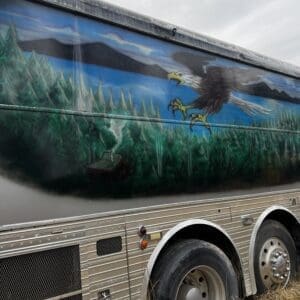 Silver trailer side painted with a large eagle soaring above a pine forest and blue lake, with its wheels visible nearby in dry grass.