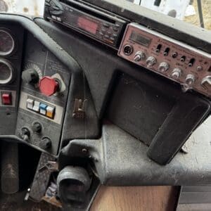 Dusty, aged vehicle dashboard with an old radio and control panel, showing worn buttons, gauges, and a large red emergency knob near the steering area.