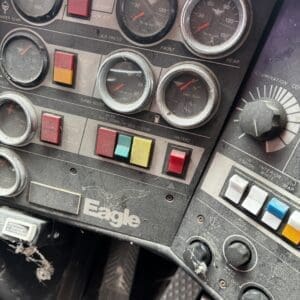 Close-up of a dusty aircraft cockpit panel: multiple round gauges, colored switches, and an Eagle logo.