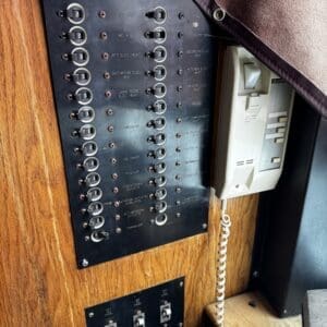 Wood-paneled wall with a large black control panel filled with rows of labeled toggle switches next to a beige wall-mounted intercom with a coiled cord
