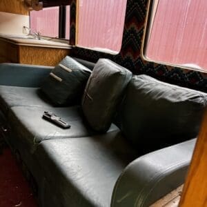 Teal leather couch inside a vintage camper, with pillows and a remote on the seat, next to a small kitchen area and red exterior outside the window.