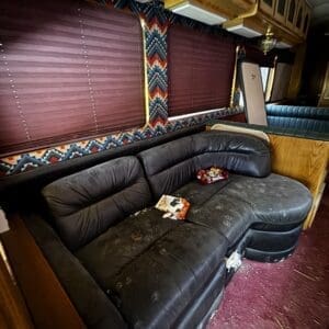 Interior of an old coach bus or RV: black leather sectional sofa, burgundy window blinds, patterned valances, wood cabinets, and scattered debris on a maroon floor.