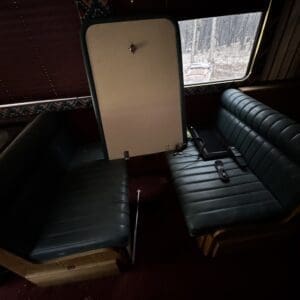 Dark-green vinyl bench seating faces each other with a fold-down white tabletop mounted between them inside a dim bus or camper interior; window shows a forest scene outside.