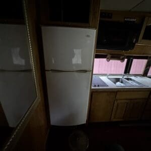 Dimly lit RV kitchen with a white double-door refrigerator between wooden cabinets and a double sink along the counter.