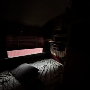 Dimly lit RV bunk bed with gray sheets and a dark pillow, window with closed blinds and overhead storage.