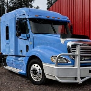 Blue semi-truck with a large silver bull bar parked on a gravel lot beside a red building.