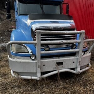 Front view of a blue Freightliner semi-truck with a metal grille guard, parked on dry grass beside a red corrugated wall/Barn.