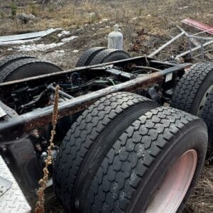 Rear view of a rusted semi-truck chassis with multiple dual tires in a bare, wooded junkyard area.