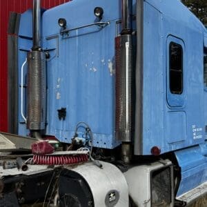 Blue semi truck cab viewed from the rear-left, with tall chrome exhaust stacks and ground hoses nearby.