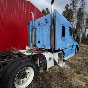 Blue semi-truck tractor with tall chrome exhaust stacks parked on dry grass beside a red metal building.