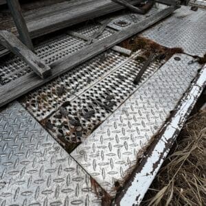Rusty diamond-plate platform with peeling paint, weathered wooden planks, and dried pine needles scattered across the surface.