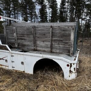 Rusty white truck bed with a weathered wooden crate on top, in a grassy field with trees in the background.