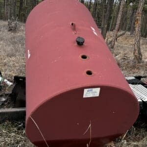 Red horizontal propane tank in a forest clearing, mounted on a wooden platform with a black fill cap and vent openings along the top.