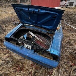 Open trunk of a blue car in a dry, grassy field, cluttered with car parts and debris; red barn and a bus visible in the background.