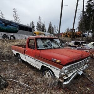 Rusted red and white vintage pickup abandoned in a grassy junkyard, with a battered chrome bumper and overcast sky, surrounded by other old cars and vehicles.