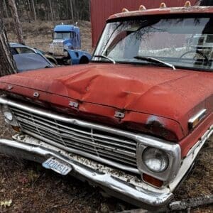 Front view of a weathered red Ford pickup with a dented hood and chrome bumper in a junkyard among abandoned vehicles.