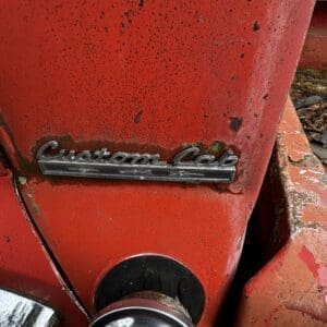 Close-up of a weathered red vintage vehicle front with a chrome 'Custom Cab' emblem and a round headlight below.