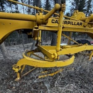 Yellow Caterpillar forestry machine frame with hydraulic arms and gears in a pine forest clearing on uneven ground.