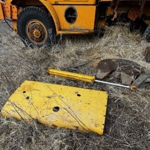 Abandoned yellow construction vehicle parts scattered in dry grass, including a cracked metal hatch and a jack beside a large tire.