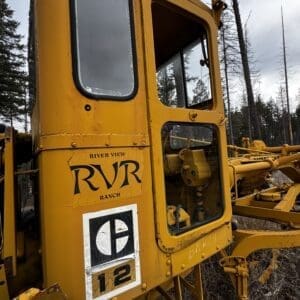 Yellow heavy machinery cab with River View Ranch (RVR) logo and a '12' label, outdoors among trees in a forested area.