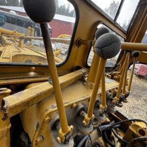 Inside the cab of a yellow construction vehicle, showing several long levers with round black knobs and exposed hydraulic hoses.