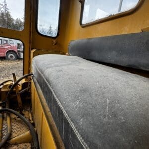 Interior of an old yellow construction vehicle with a dusty gray seat, exposed hoses, and metal diamond-plate floor; outside shows trees and a red vehicle through windows