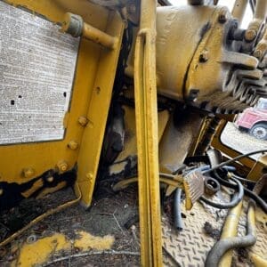 Close-up of yellow construction equipment showing a hydraulic piston, metal brackets, and dirt-covered surfaces on a diamond-plate floor.