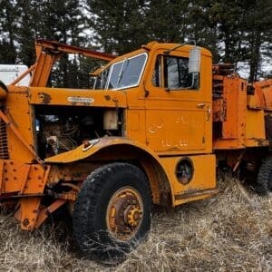 Old orange Oshkosh heavy tow truck, with exposed engine and front winch, parked in tall dry grass.