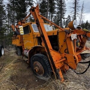 Abandoned orange tow truck with a tall crane arm in a grassy forest clearing, rusted and worn.