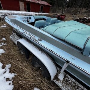 Vintage turquoise speedboat on a trailer in a rural yard with a red barn and pine trees in the background, patches of snow on the ground.