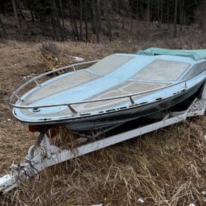 Blue vintage speedboat on a trailer parked in a dry, snow-dotted field with a forest in the background.”