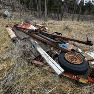 Scrap metal and a spare tire piled on a flatbed trailer in a dry grassy clearing with a forest of tall pine trees behind.