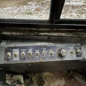 Old vehicle dashboard with weathered metal panel and labeled switches for wheels, lamps, chime, idle, heat, AC, and heater adjustments seen at an outdoor windowed cabin edge.