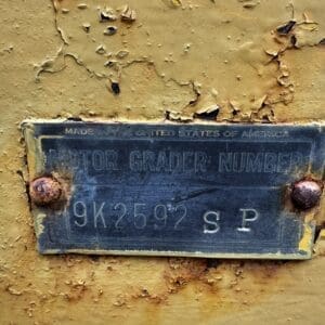 Close-up of a weathered metal ID plate on a yellow, rusting surface; reads 'Motor Grader Number 9K2597 SP' (Made in USA).